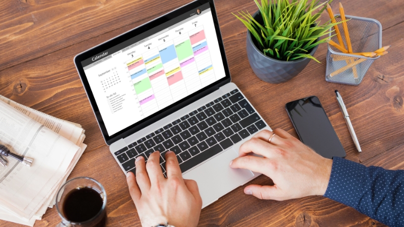 A person works on a laptop showing a colorful social media holidays calendar on a wooden desk