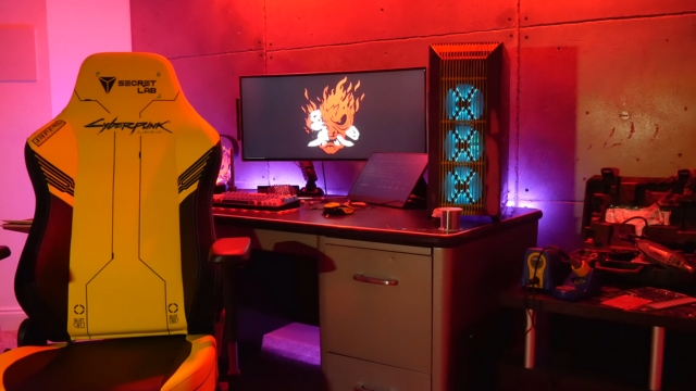 A yellow Cyberpunk 2077 chair sits in a neon-lit setup with a monitor and tower glowing in red and blue
