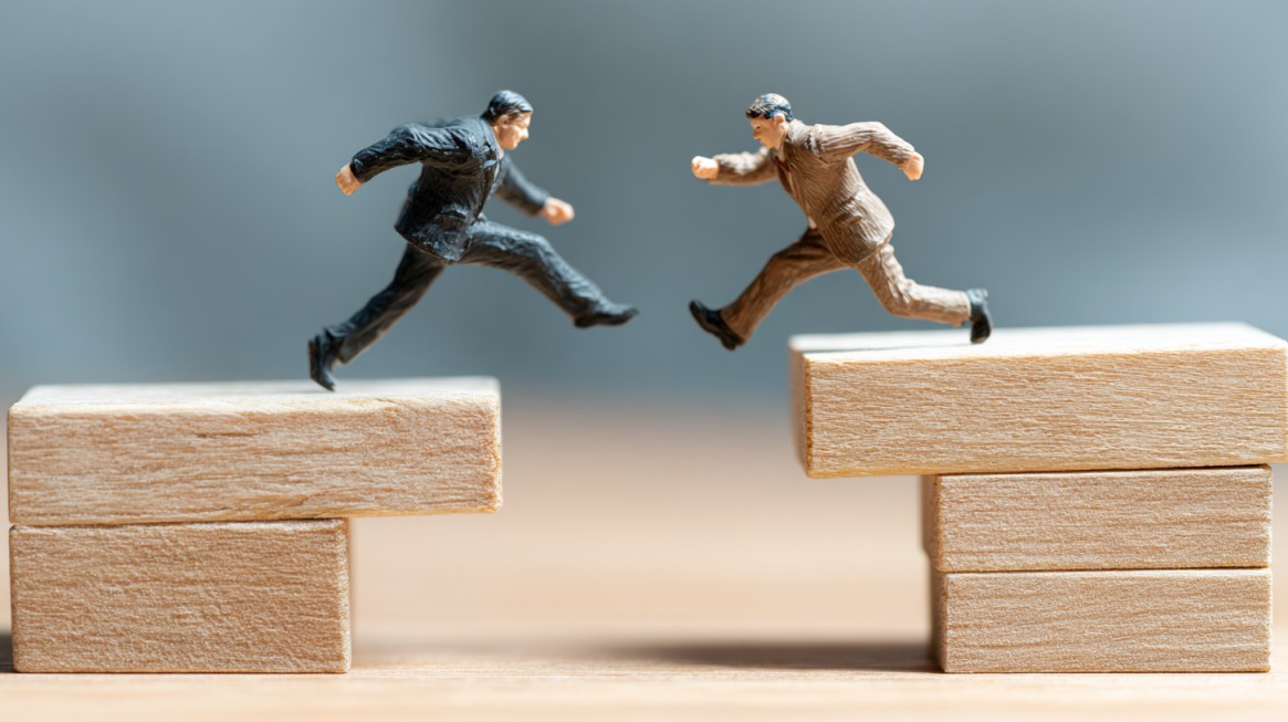 Two miniature business figures leaping across wooden blocks toward each other, symbolizing competition and agility
