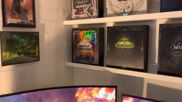 Collector’s edition WoW boxes line the shelves above a curved monitor showing an in-game scene