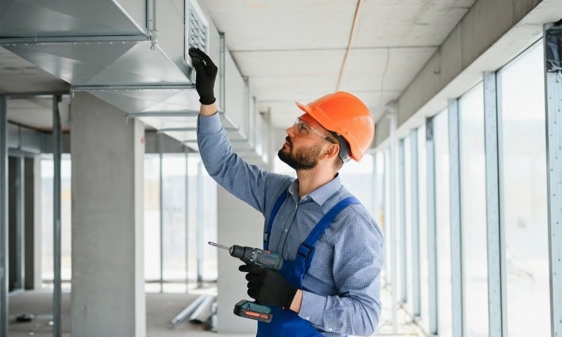 A man in an orange hard hat and overalls holds a drill, repairing HVAC system