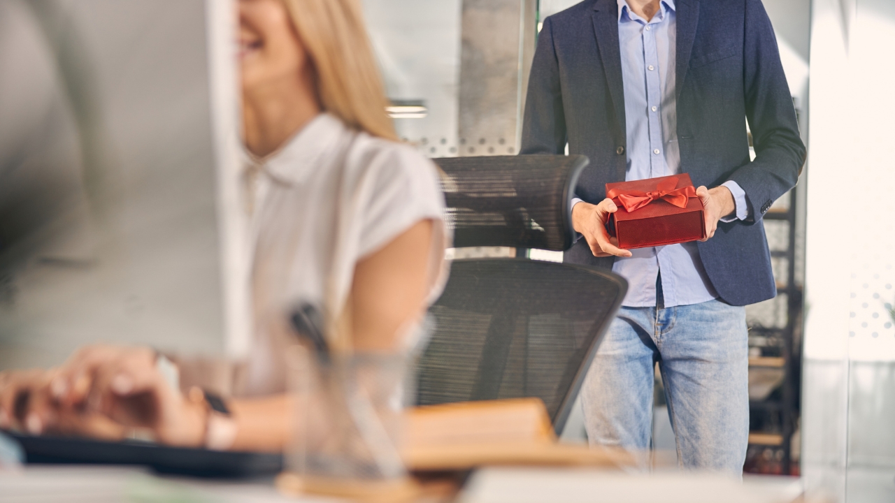 Man holds a gift box behind a woman at an office desk